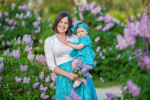 Happy mother mom with daughter enjoying time on a awesome place between lilac syringe bush.Young ladies with basket full of flowers dressed in jeans and stylish shirts relaxing in a cloud of scent.