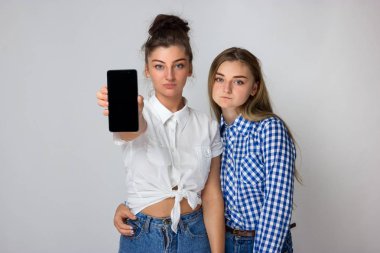 Gri arka plan üzerinde duran ve kameraya bakarak boş smartphone ekran gösterilen mutlu iki genç kız portresi