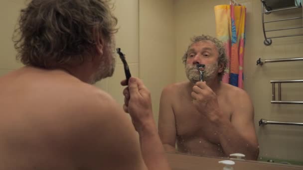 Muž v koupelně před zrcadlem oholí vousy s břitvou. — Stock video