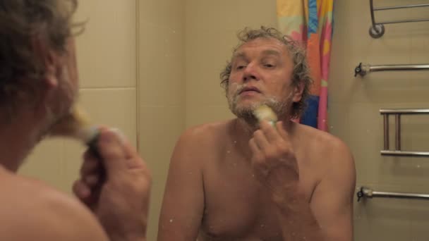 A handsome adult man looks in the mirror and shaves his beard. — Stock Video