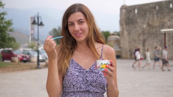 Милая молодая женщина идёт по улице и ест ложечным мороженым — стоковое видео
