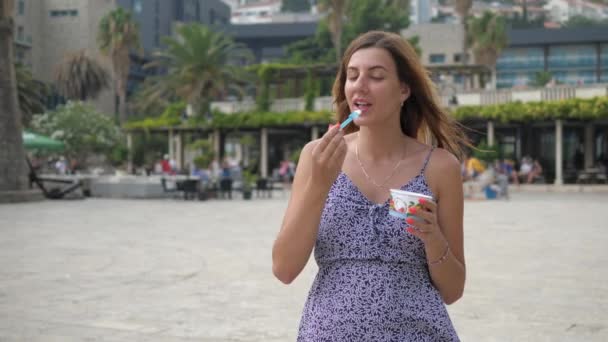 Bella giovane donna cammina lungo la strada e mangiare con un cucchiaio di gelato — Video Stock