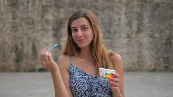 Carino giovane donna mangiare con un cucchiaio da una tazza di gelato per strada — Video Stock