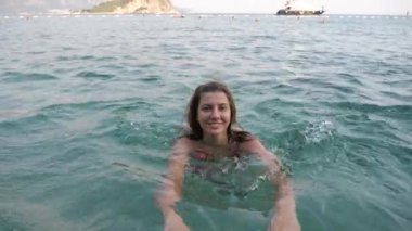 Genç bir kadın denizde, iplik ve kameraya bakarak eğleniyor yüzüyor