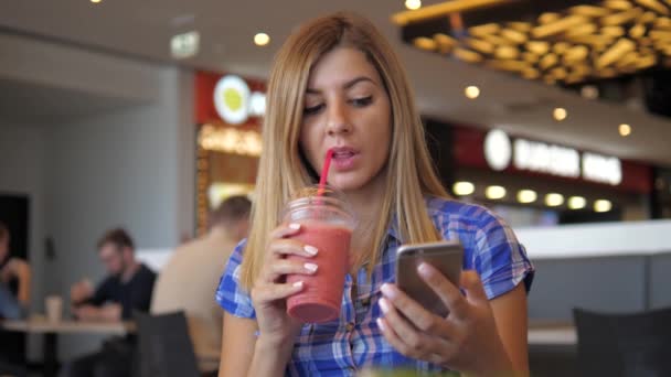 Jovem bonita mulher bebe suco do tubo para o café, usando um smartphone — Vídeo de Stock