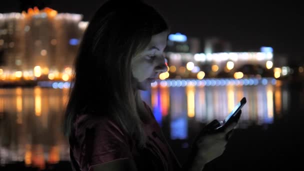 На Ночной улице женщина работает на смартфоне. На заднем плане - ночные городские огни — стоковое видео
