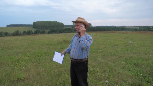 Anciano granjero en sombrero de vaquero de pie en el campo, habla por teléfono — Vídeos de Stock
