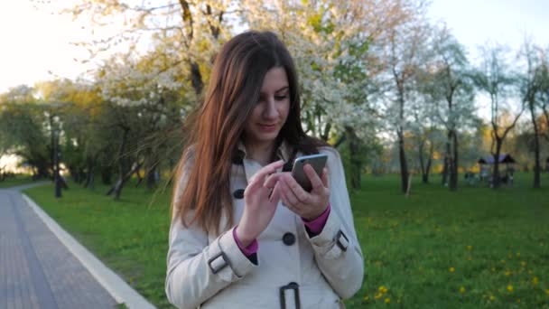 Linda e linda garota morena de casaco usa o telefone de pé em um parque — Vídeo de Stock