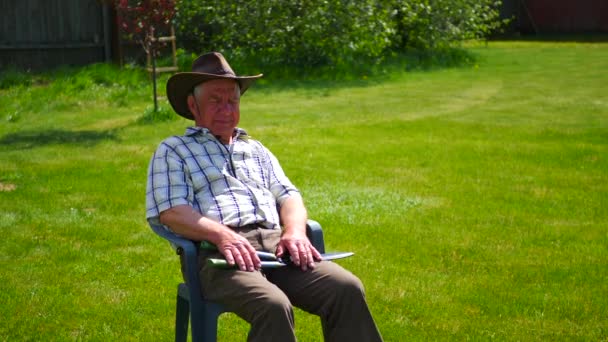Starý člověk je unavený a sedí v křesle s kloboukem na zeleném trávníku. — Stock video