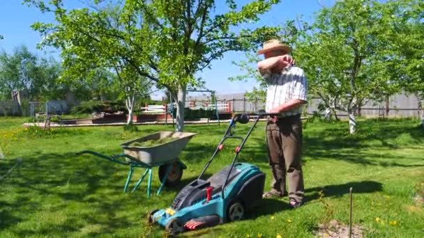 Um homem velho está de pé com um cortador de grama em um belo jardim . — Vídeo de Stock