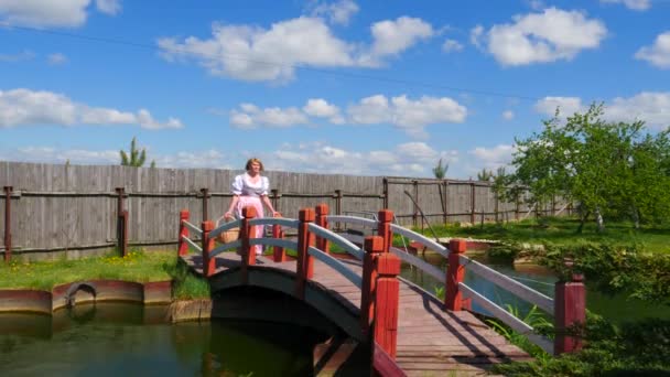 Žena v národních krojích jde s košíkem na mostě přes rybník. — Stock video