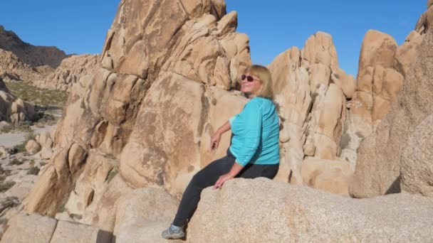 Mujer activa se sienta en lo alto del borde de la roca, regocijando la vista y sonriendo — Vídeos de Stock
