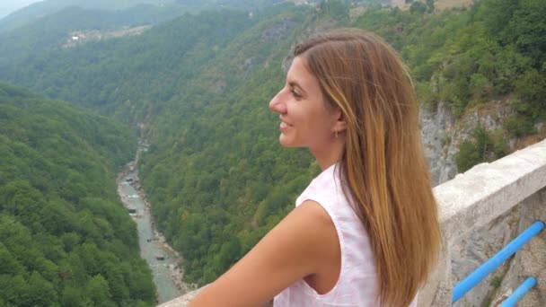 Mujer joven mirando las montañas de pie en un puente sobre un cañón profundo — Vídeos de Stock