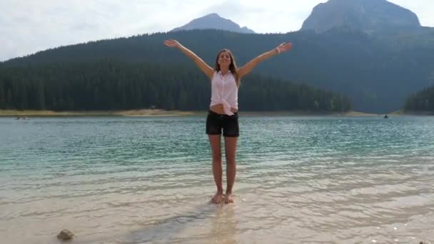 山の湖で石の上に立っている女性は、側面まで手を伸ばしてください。 — ストック動画
