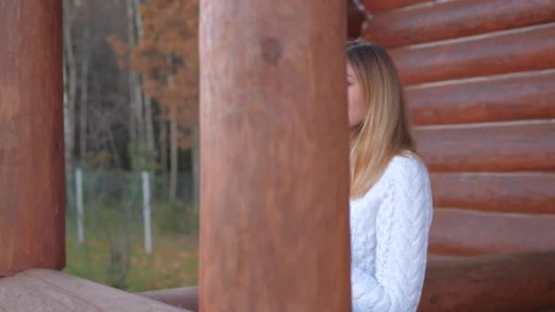 Mujer en suéter blanco con taza de té va en la terraza de una casa de madera — Vídeo de stock
