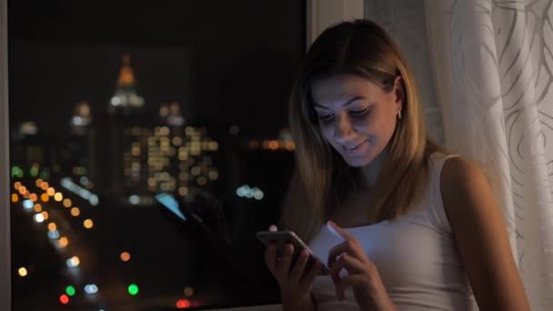 Красива жінка, стоячи біля вікна з видом на ніч і використовує смартфон — стокове відео