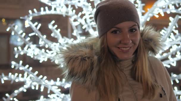 Молодая женщина в теплой одежде улыбается, стоя посреди рождественских иллюминаций — стоковое видео