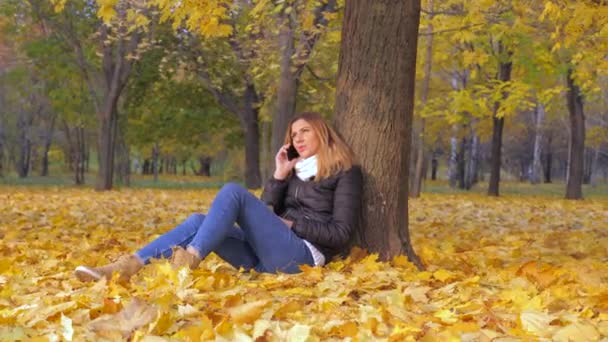 Женщина сидит спиной к дереву в жёлтых осенних листьях, разговаривает со смартфоном — стоковое видео
