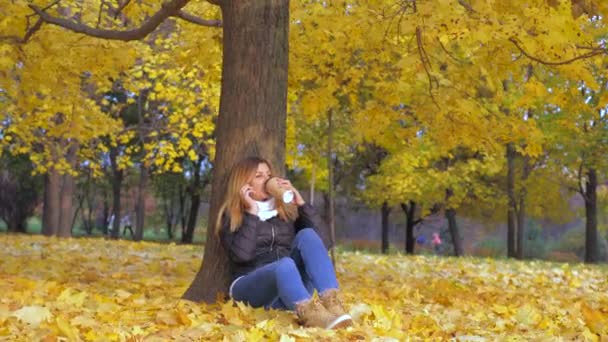 Frau sitzt mit dem Rücken zum Baum in gelben Herbstblättern und spricht Smartphone — Stockvideo