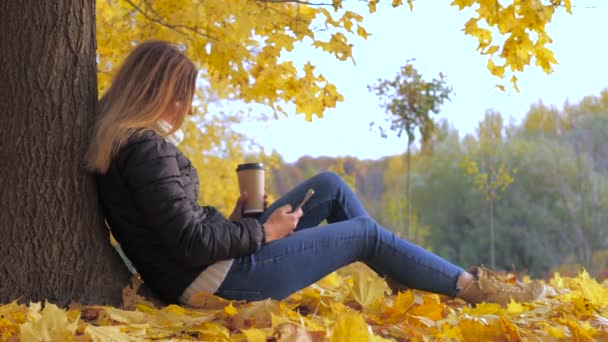 Mulher sentada de volta à árvore na folhagem de outono amarelo, usa o telefone 4k — Vídeo de Stock