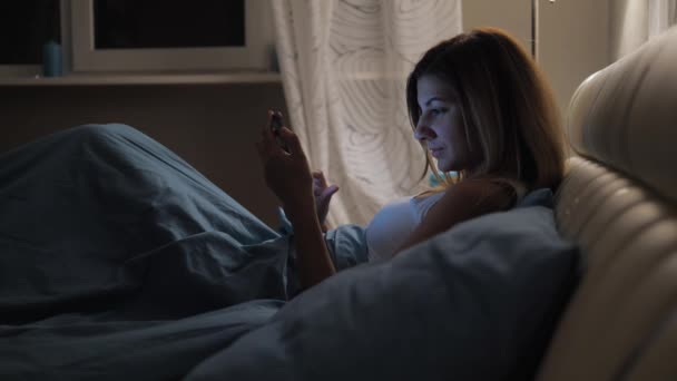 Mulher na cama usa aplicativo no smartphone antes de ir dormir Olhando para fotos — Vídeo de Stock