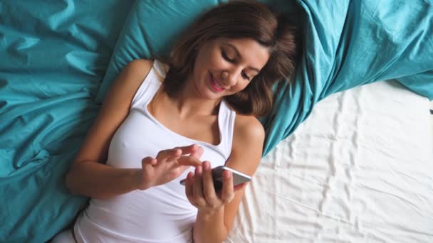 Vista dall'alto della donna abbronzata sexy al mattino sdraiata su un letto utilizza un'app per smartphone — Video Stock