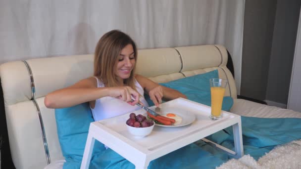 Женщина ест сосиски на завтрак с яичницей на столе, лежа в постели — стоковое видео