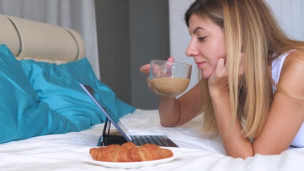 Giovane donna con computer portatile mangiare croissant a letto a casa e bere caffè . — Video Stock