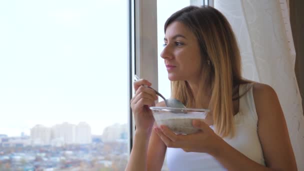 Vrouw eet ontbijtgranen met melk uit kom staande op venster en op zoek naar buiten — Stockvideo