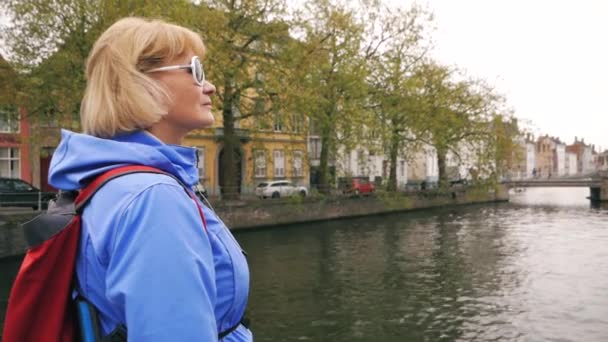 Touristin steht am Flusskanal, auf dem in der alten Steinstadt ein Ausflugsboot schwimmt — Stockvideo
