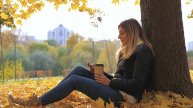 Sarı sonbahar yaprakları, ağacında yakın oturan bir Bayan Apps ve kahve içme kullanır