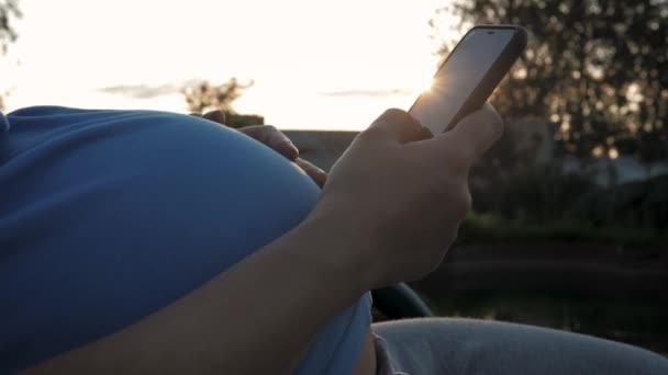 Mujer embarazada con gran abdomen utiliza aplicaciones de teléfonos inteligentes sentado al aire libre al atardecer — Vídeo de stock