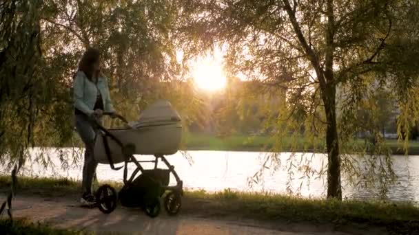 Mujer camina por la calle con un cochecito de bebé contra el sol al atardecer — Vídeos de Stock