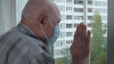 Koruyucu tıbbi maskeli yaşlı adam karantina sırasında pencereden dışarı bakar.