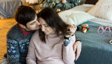 A man in a New Year's patterned sweater gently hugs his wife by the shoulders in the bedroom decorated for the New Year sitting near the bed. The man smiles, the woman rested her head on his shoulder.