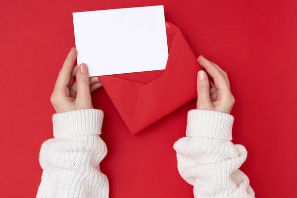 Kobiece ręce trzymające pustą pocztówkę i czerwoną kopertę na czerwonym tle. Koncepcja świąteczna — Zdjęcie stockowe