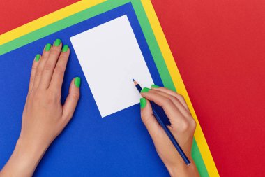 Çok renkli bir arka planda yeşil tırnaklı, boş bir kartpostalın üzerinde mavi kalem tutan kadın ellerini kapat.