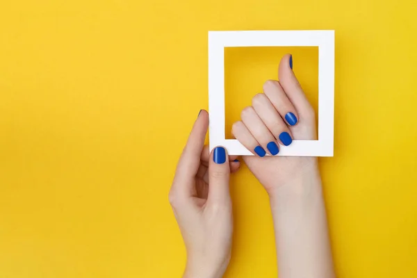 Female hands with manicure in trendy classic blue color holding white frame on the yellow background — Stock Photo, Image