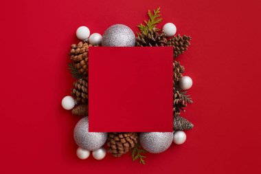 Köknar kozalakları ve kırmızı zemin üzerinde beyaz ve gümüş parıldayan toplar kompozisyonu ortasında boş bir kırmızı tebrik kartı düz yatıyordu. Noel konsepti