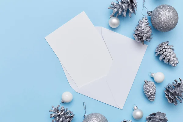 Zarfın içinde gümüş köknar konileri ve mavi arka planda beyaz ve gümüş toplarla çevrili boş bir beyaz tebrik kartının düz kompozisyonu vardı. Noel konsepti — Stok fotoğraf