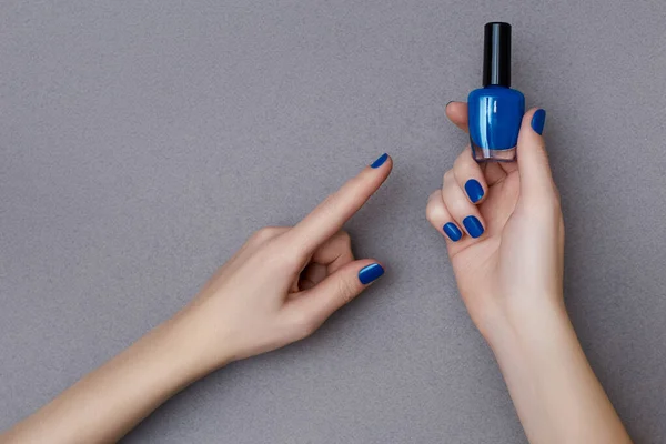 Weibliche Hände mit Maniküre in trendigen klassischen blauen Farbe hält eine Flasche Nagellack auf dem grauen Hintergrund — Stockfoto