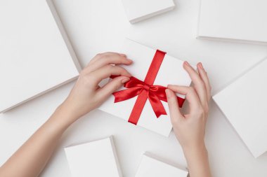 Kırmızı kurdeleli beyaz bir hediye kutusu ve beyaz arka planda beyaz kutularla çevrili bir yayı bağlayan kadın eli.