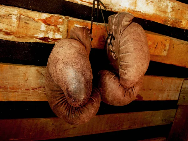Boxing gloves on the wall. Old, vintage pair of leather mittens hangs on the wood wall. Red colors and soft lights. Gloves of retired boxer and fighter. It hangs on a nail in a barn or rustic house. — Stock Photo, Image