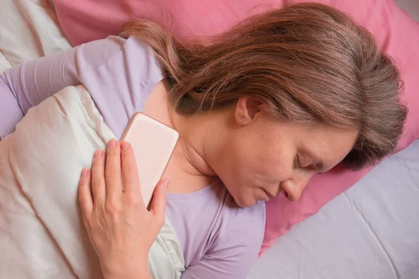 女人睡在床上带着手机 40岁的中年妇女躺在枕头上 手里拿着智能手机 社交网络或网瘾 — 图库照片