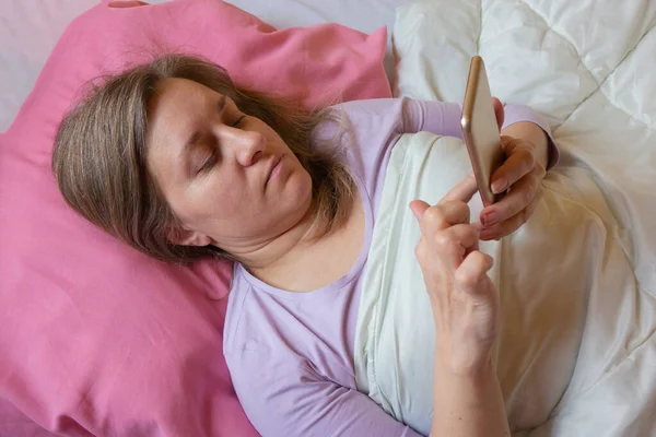 女人在床上用手机 40岁的中年妇女躺在枕头上 用智能手机发短信 社交网络或网瘾 — 图库照片