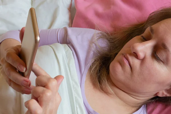 女人在床上用手机 40岁的中年妇女躺在枕头上 用智能手机发短信 社交网络或网瘾 — 图库照片