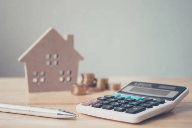 Tahta ev, bozuk para ve tahta masa üzerine kalem yığılı hesap makinesi. Emlak yatırımı ve konut kredisi finansal konsepti