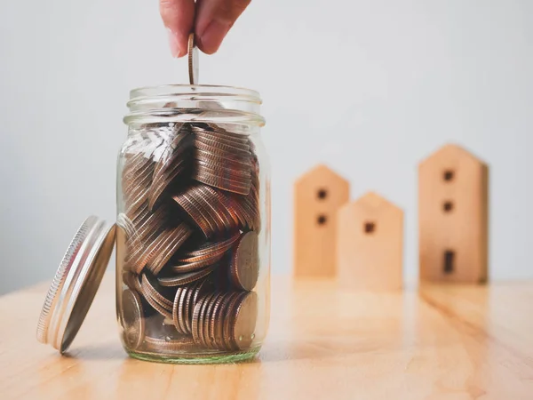 Property investment real estate and house mortgage financial concept. Hand putting money coins in jar with wooden home on table