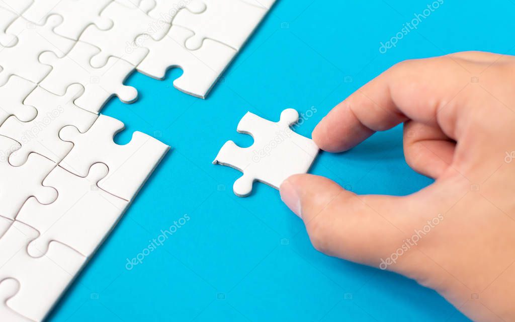 Hand putting piece of white jigsaw puzzle on blue background. Team business success partnership or teamwork.