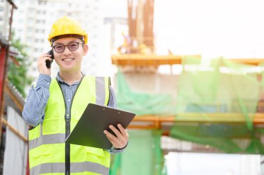 Genç Asyalı mühendisler inşaat alanında çalışıyorlar. Sarı kask ve üniforma giyin. İletişim ve evrak işleri için taşınabilir radyo alıcısı.